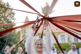 At Château de La Salle, la Pacaudière, the bride stands outside with eyes closed, holding scissors overhead to cut ribbons during the bouquet game, surrounded by eager guests hoping to win the prize.